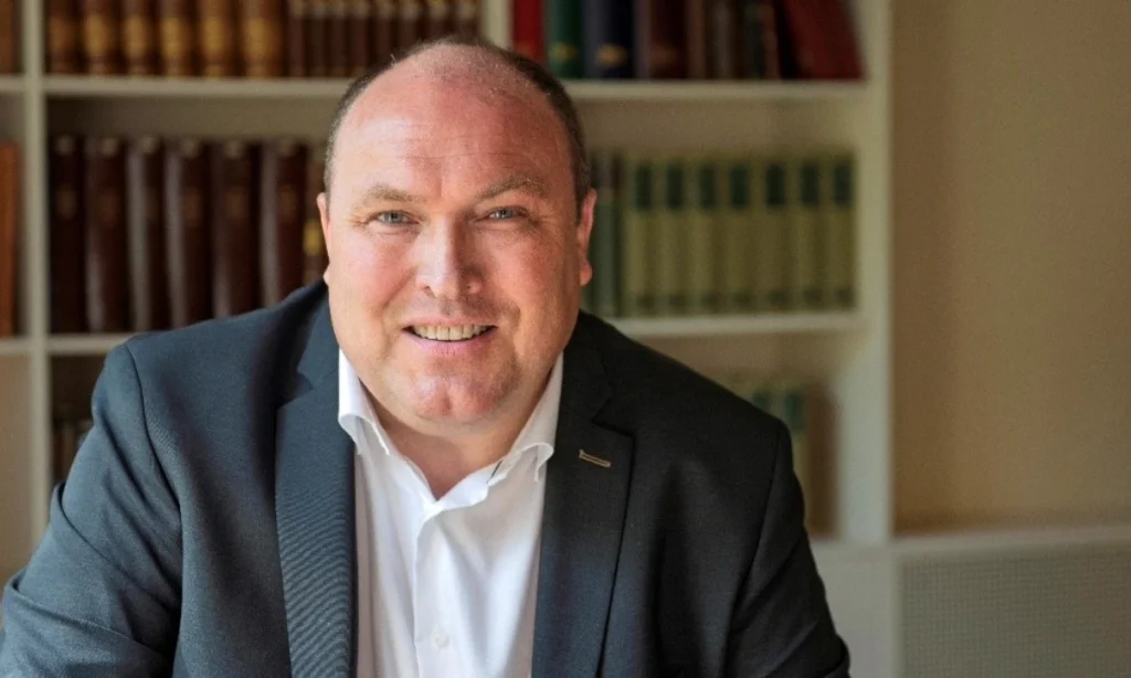 Biskop Thomas Reinholdt Rasmussen i jakkesæt smiler foran en bogreol, klar til at holde grundlovstale.